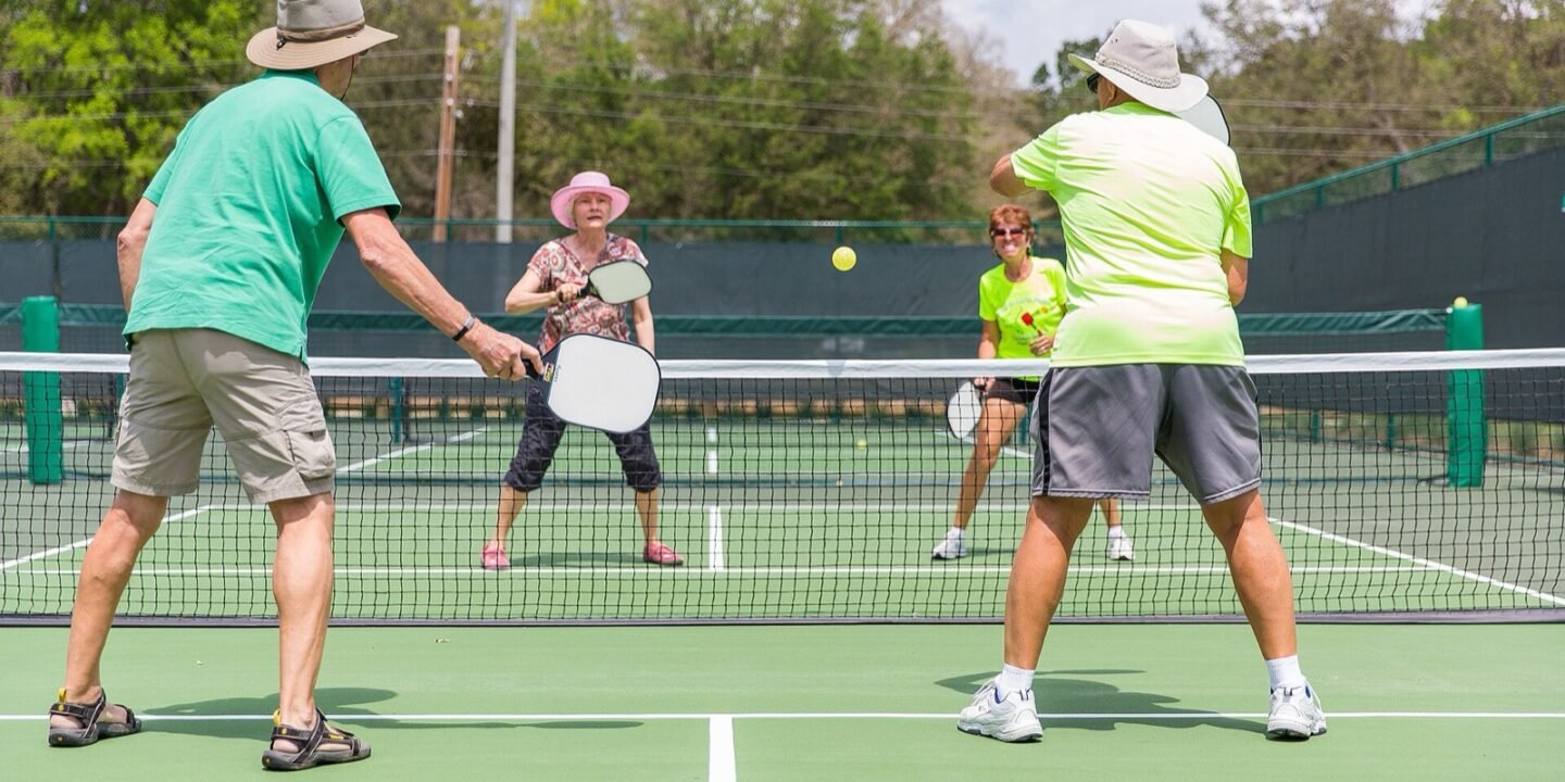 four players in sun hats trading shots at the net on an outdoor pickleball court